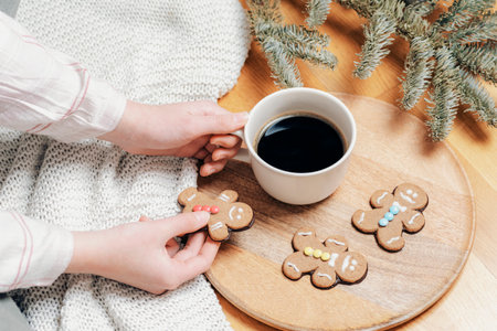 Top view of hands holding a coffee cup with gingerbread cookies on a wooden tray and festive pine branches on a knitted blanket.の写真素材