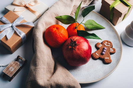 Festive still life with pomegranate, oranges, gingerbread cookies, and wrapped gifts on a white...