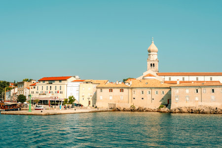 Scenic view of a coastal town with stone buildings, a church tower, and calm blue water under a clear sky, showcasing Mediterranean architecture. Krk island, Croatia.の写真素材