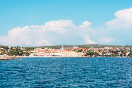 Panoramic view of a Mediterranean coastal town with red roofs, blue sea, and green hills in the background on a sunny day. Krk island, Croatia.の写真素材