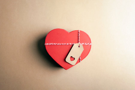 Red heart-shaped box with a kraft tag on a beige background, representing love and thoughtful gifting. Valentines day concept. Top view, flat lay.の写真素材
