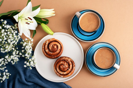 Cinnamon rolls on a white plate with two cups of coffee in blue ceramic cups, white lilies, and gypsophila flowers arranged on a beige background with a dark blue fabric. Top view, flat lay.の写真素材