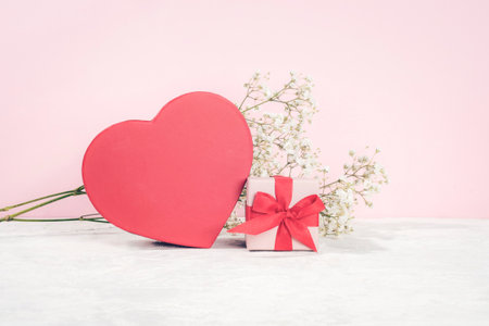 Red heart-shaped box and a small gift box tied with a red ribbon next to babys breath flowers on a light pink background. Valentines day concept.の写真素材