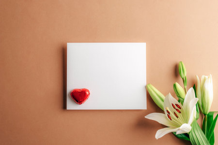 White lily with blank card and heart-shaped candy on a beige background, symbolizing purity and love. Valentines day concept. Top view, flat lay, mockup.の写真素材