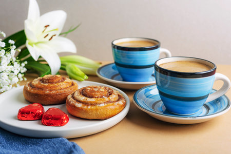 Two blue coffee cups with cinnamon rolls and white lilies on a beige background, symbolizing relaxation and romantic moments. Valentines day concept.の写真素材