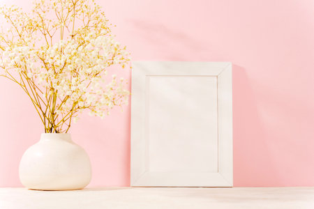 White vase with baby's breath flowers and blank picture frame against a pink wall, symbolizing delicate beauty and elegance. Spring concept, mockup.の写真素材