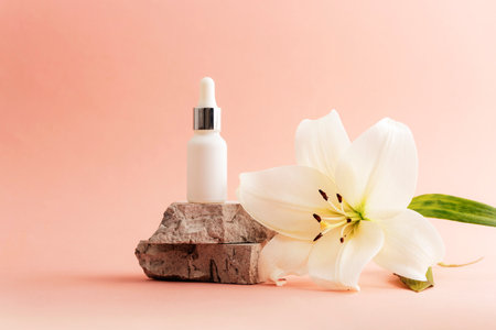 White cosmetic serum dropper bottle with a white lily flower on a stone against a pink background. Natural cosmetics concept. Closeup.の写真素材
