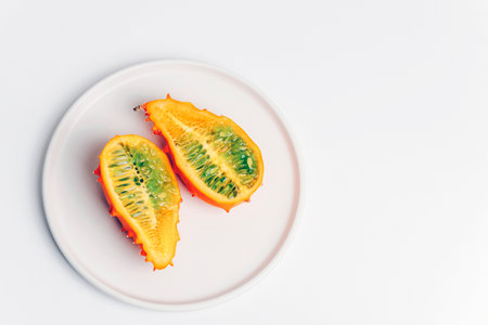 Two halves of ripe kiwano fruit with orange skin and green pulp on a white plate on white background.の写真素材