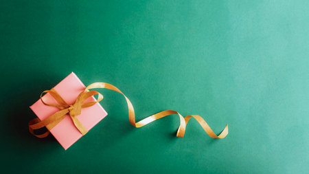 Pink gift box with a shiny golden ribbon on a deep green background. Holiday greeting card. Top view, flat lay, copy space.の写真素材
