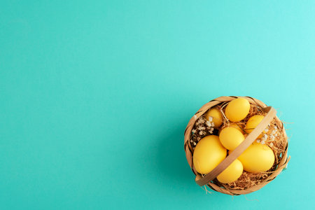 Wicker basket filled with yellow Easter eggs, straw, and dried flowers on a pastel turquoise...