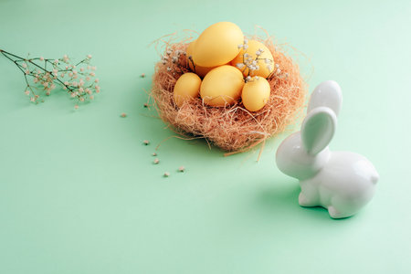 Easter nest with yellow eggs, dried flowers, and a white ceramic bunny on a pastel green background,...