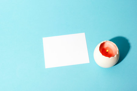 White blank card and burning candle in eggshell-shaped holder on pastel blue background. Easter...