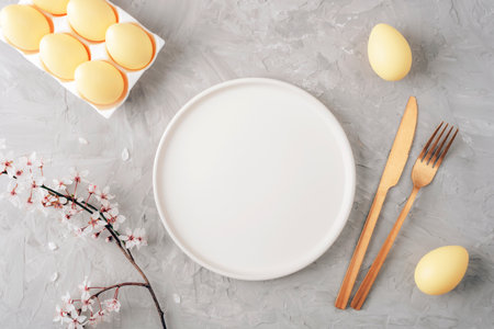 Empty white round plate with golden cutlery, yellow eggs and cherry blossom on grey background....
