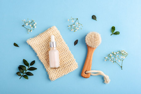 Cosmetic serum bottle with wooden dry brush, delicate flowers and green leaves arranged on light blue background in flat lay compositionの写真素材
