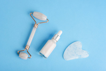 Skincare tools including facial roller, serum dropper bottle and gua sha stone on blue background in minimal flat lay compositionの写真素材