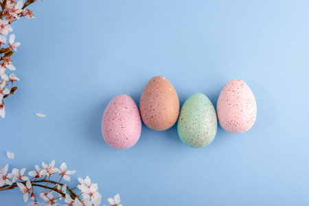 Four pastel speckled Easter eggs on blue background with cherry blossom branches and scattered petals. Easter concept. Top view, flat layの写真素材