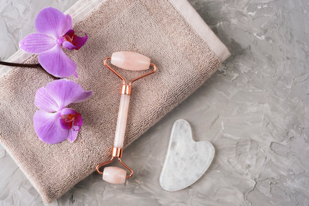Skincare roller and gua sha stone with orchid flowers on towel on grey background. Flat lay, spa conceptの写真素材