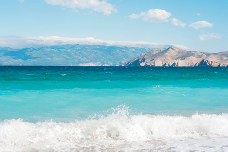 Blue ocean waves crashing on sandy beach with mountains in background.の写真素材