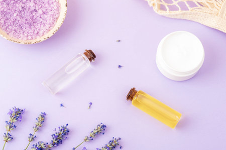 Lavender bath salt in bowl, cream jar, lavender flowers, essential oils on purple background. Natural herbal medicine, aromatherapy concept. Top view, flat layの写真素材