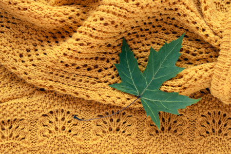 Green maple leaf lying on warm knitted yellow fabric creating cozy autumn seasonal background. Top...