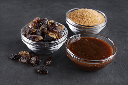 Caramelized sugar, brown sugar cane and liquid caramel in glass bowls on dark concrete backgroundの写真素材
