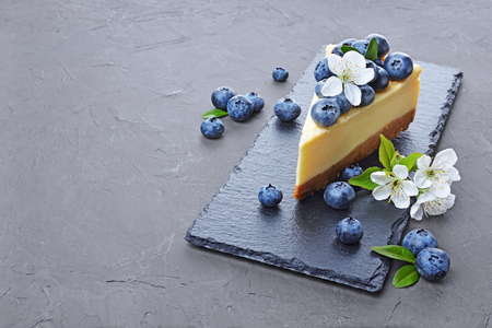 Close-up slice of delicious homemade cheesecake with fresh blueberries and spring flowers on black slate board on gray concrete backgroundの写真素材