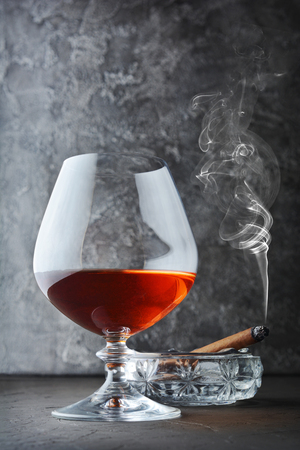 Strong alcoholic drink cognac in sniffer glass with smoking cigar in crystal ashtray on gray concrete backgroundの写真素材