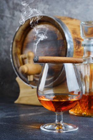 Strong alcoholic drink cognac in sniffer glass with smoking cigar, crystal decanter and vintage wooden barrel in cellar on gray concrete backgroundの写真素材