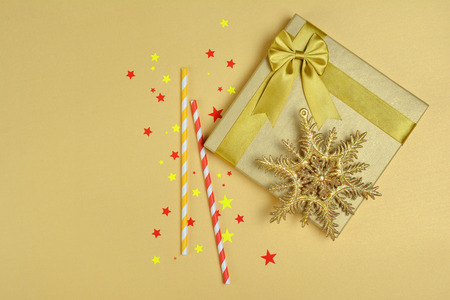 Shiny classic gift box with satin bow, cocktail straws and glittering christmas tree toy snowflake as attributes of party on golden backgroundの写真素材