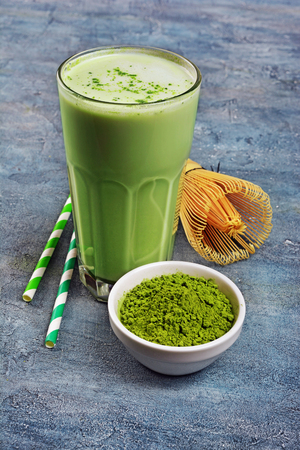 Organic green matcha tea latte in glass and powder in white bowl with bamboo whisk and cocktail straw on blue concrete backgroundの写真素材