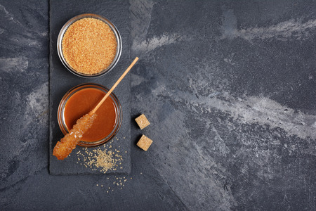 Top view on homemade sweet caramel sauce in glass bowl and brown cane sugar with sugar crystal stick on black slate board and marble backgroundの写真素材