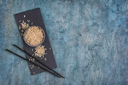 Bowl of uncooked brown long grain rice with wooden chinese chopsticks on black slate board and blue concrete backgroundの写真素材