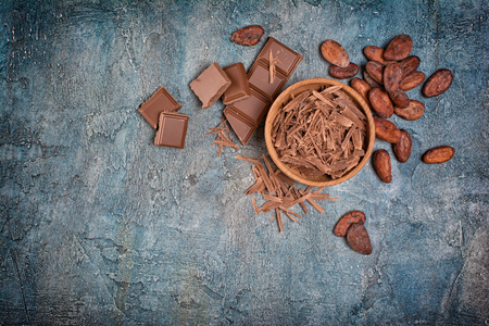 Top view of chocolate chips and pieces of bars with cocoa beans for confectionery on blue concrete background with copy spaceの写真素材