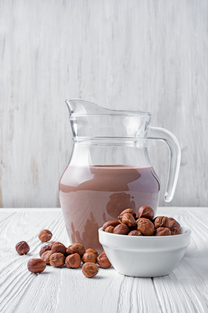 Healthy natural chocolate milk in glass jug with hazelnuts for breakfast on white wooden backgroundの写真素材