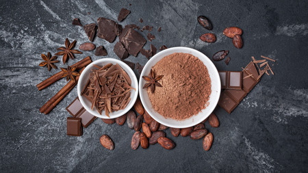 Top view of cocoa powder and chocolate chips in white bowls and whole cocoa beans with spices as ingredient for confectionery on black marble backgroundの写真素材