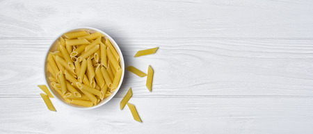 Top view of wide textured white wooden banner background with Italian raw penne rigate pasta in bowl with copy spaceの写真素材