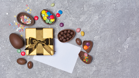 Top view on chocolate traditional Easter eggs and golden gift box with white card and bright colorful dragee, sugar sprinkles on gray concrete backgroundの写真素材