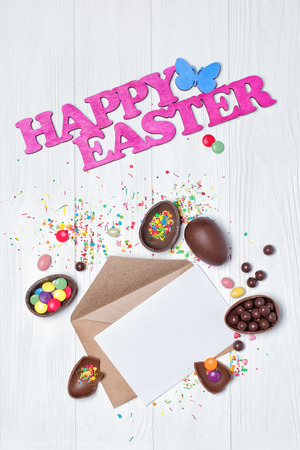 Top view on mockup of blank white card in kraft envelope with pink text of happy Easter and chocolate traditional Easter eggs on white wooden backgroundの写真素材