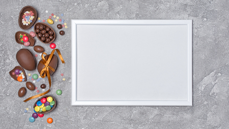 Top view on mockup of blank white frame traditional easter sweets as chocolate eggs and bright colorful sugar sprinkles or confetti on gray concrete backgroundの写真素材