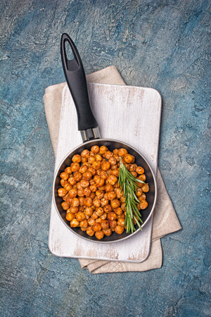 Roasted salty and spicy chickpeas in frying pan with rosemary on white wooden board and concrete background with copy spaceの写真素材