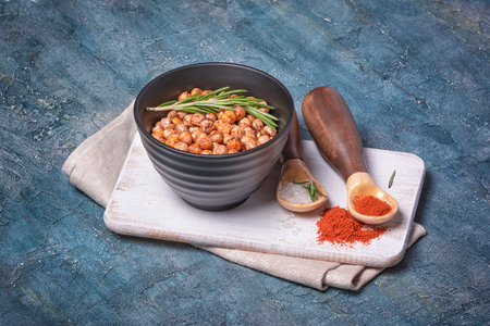 Roasted spicy chickpeas in black bowl with rosemary, sea salt and paprika in spoons on white wooden board and blue concrete backgroundの写真素材
