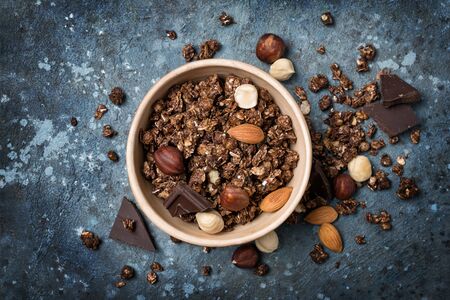 Top view of oatmeal or granola with pieces of dark chocolate, almond and hazelnut in bowl for nutritious breakfast on blue concrete backgroundの写真素材