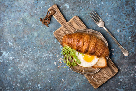 Top view of croissant sandwich with fried egg and arugula leaves on vintage silver plate and blue concrete background with copy spaceの写真素材
