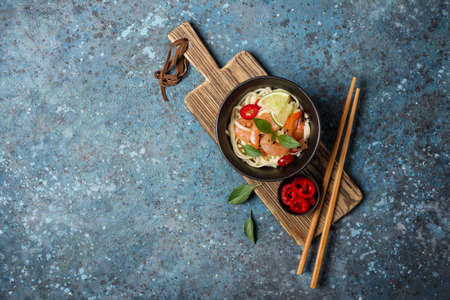 Top view of bowl with japanese udon noodles, spicy shrimps, lime and black sesame seeds on wooden board and blue concrete background with copy spaceの写真素材