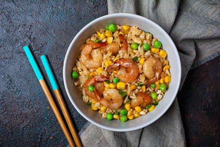 Top view of bowl with chinese rice, tasty prawns, yellow corn, chicken egg, green onion and wooden chopsticks on dark concrete backgroundの写真素材