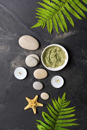 Spa and beauty concept. Top view of grey sea stones, cosmetic clay and aroma candles on black marble...