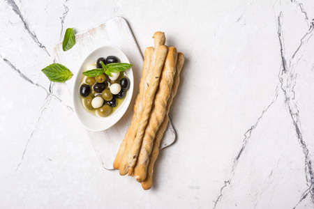 Top view of grissini with basil spice, green and black olives in oil, mozzarella on white wooden board and marble background with copy spaceの写真素材
