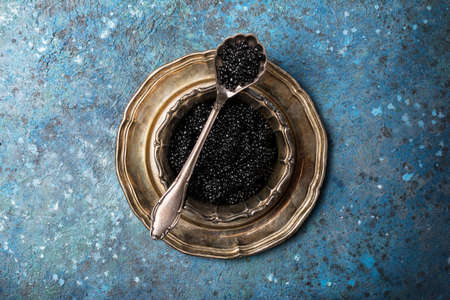 Top view of bowl with appetizing black caviar and vintage silver spoon on blue concrete background