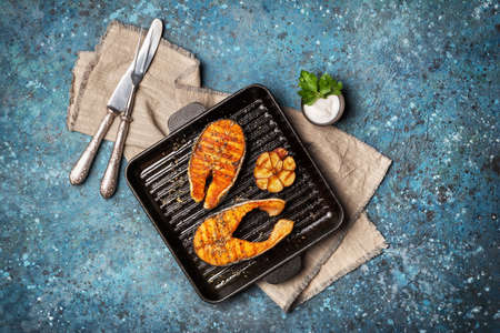 Top view of grilled red fish salmon steak on cast iron skillet or pan with spice, fried garlic and tasty sauce on blue concrete backgroundの写真素材