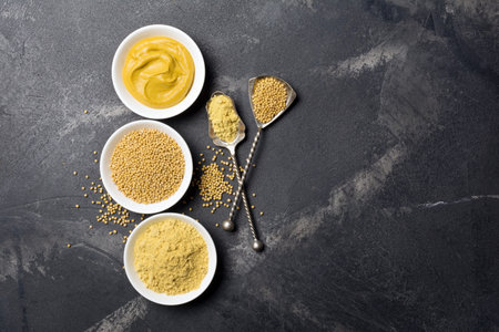Top view of mustard set in white bowls for traditional homemade sauce and spicy food with vintage spoons on black marble background with copy spaceの写真素材